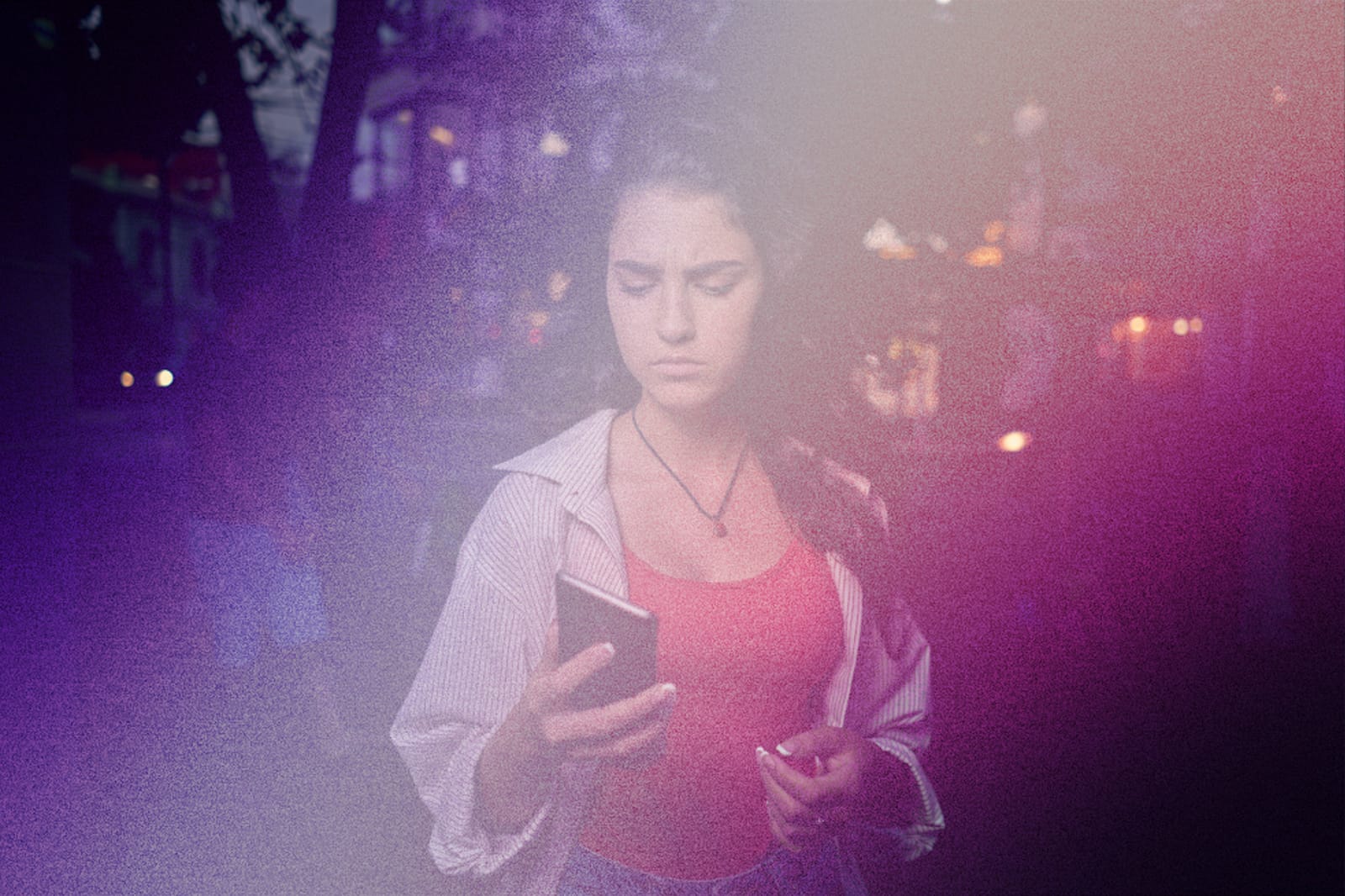 A young woman standing on a city street at night, looking down at her phone