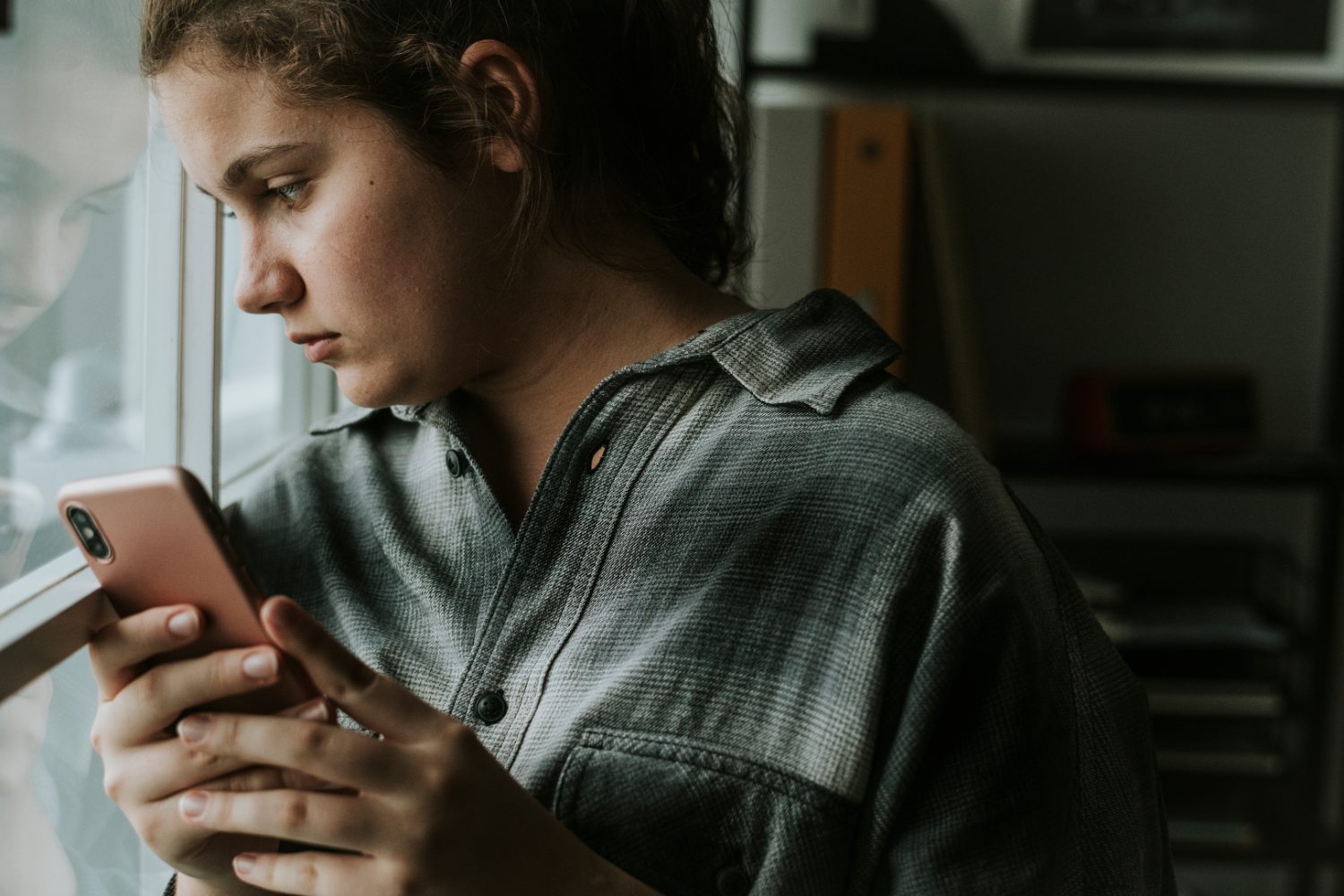 A teenage girl looks at her phone by a bright window